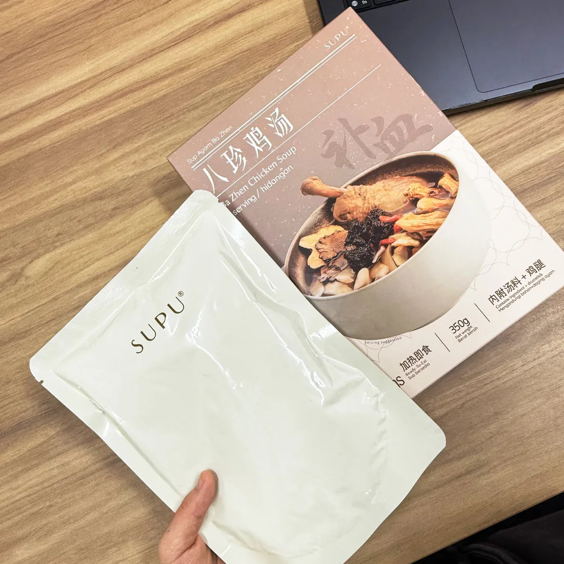 手拿 SUPU 汤包，旁边是八珍鸡汤盒，桌上有笔电